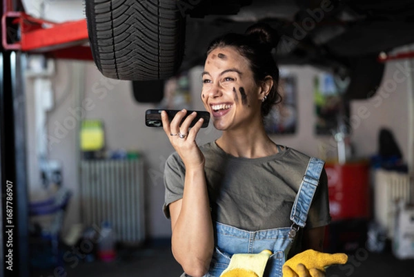 Obraz A laughing mechanic enjoys a phone conversation while working on a car in her tools-filled garage, radiating positivity.