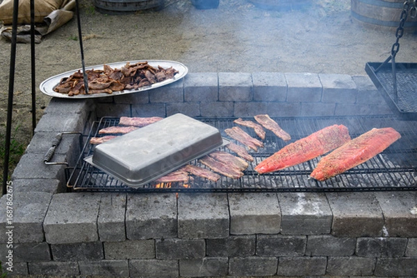 Obraz King salmon filets with teriyaki sauce grilling on an outdoor barbeque pit, as part of luau dinner preparations, stainless steel platter of cooked Korean style beef keeping warm
