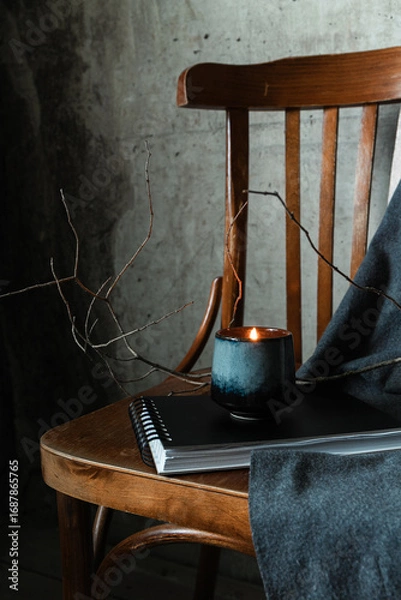 Fototapeta Lit candle in ceramic cup on notebook with wooden chair and gray fabric. Cozy minimal still life in wabi-sabi mood.