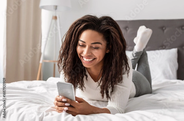 Fototapeta Mobile Phone User. Portrait of happy African American woman lying in bed and smiling while reading text message from someone, holding and using smartphone, chatting with lover or browsing social media