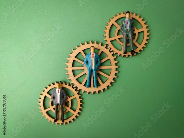 Fototapeta The image depicts a small figurine of a businessman standing on three interconnected wooden gears, set against a plain green background
