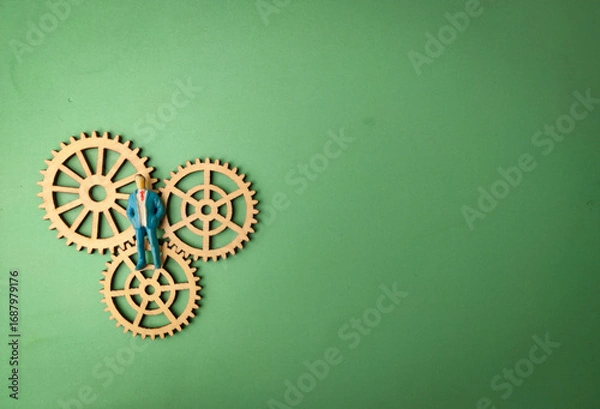 Fototapeta The image depicts a miniature figurine of a person in a suit standing on three wooden gears, arranged in a meshing configuration, all set against a plain green background