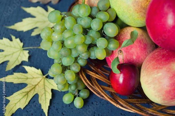 Fototapeta Harvest of ripe fruits - apples, grapes, plums, pears in a large wooden plate on a black background, decorated with autumn maple leaves. Harvest concept