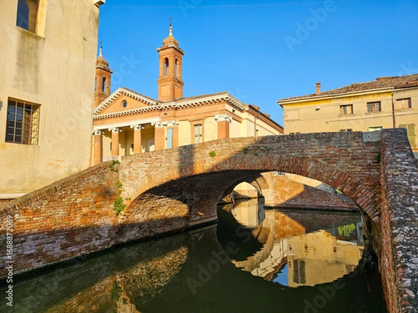 Fototapeta San Pietro church and brick arch bridge over a canal in Comacchio, Emilia-Romagna, Italy, with reflection in calm water