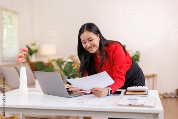Fototapeta Portrait of a asian woman using laptop work on documents and a tablet while sitting on a desk at home
