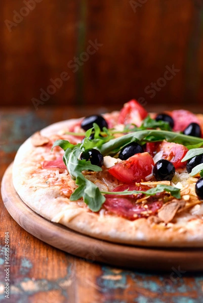 Fototapeta Pizza with salami, tomatoes and mozzarella cheese, black olives and arugula. Rustic wooden background. Soft focus.	