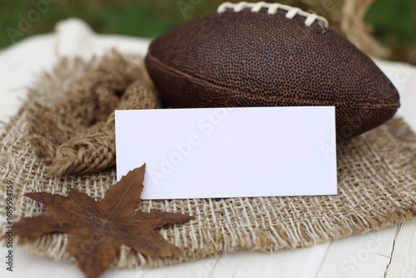 Fototapeta Blank white horizontal card mockup on fabric backing with rugby ball and dried maple leaves, soft natural light.