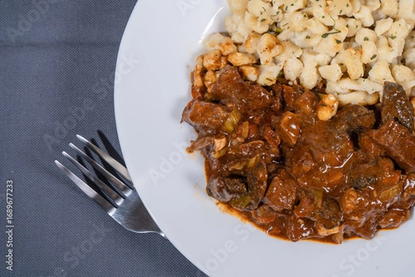 Fototapeta Delicious hungarian goulash with spaetzle on white plate resting on gray tablecloth with fork
