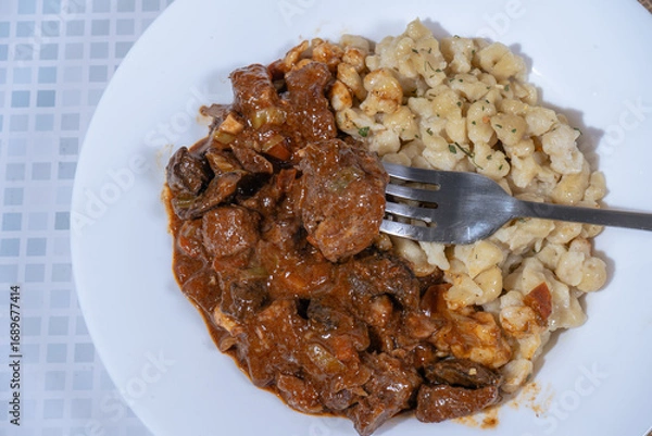 Fototapeta Fork taking a bite of beef goulash with spaetzle on white plate