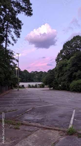 Obraz Evening view of an empty, rundown parking lot bordered by green trees with a single white cloud floating in a pink and blue sky at twilight