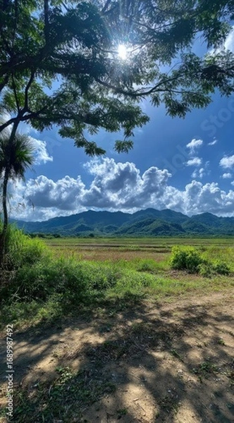 Obraz Scenic landscape of verdant fields leading to rolling mountains under a bright sky with fluffy clouds, partially framed by tree branches and foliage