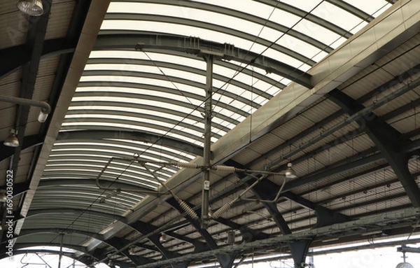 Fototapeta Low-angle view of a modern elevated train station with a curved translucent roof, dark metal beams, and visible overhead wires and lighting fixtures.