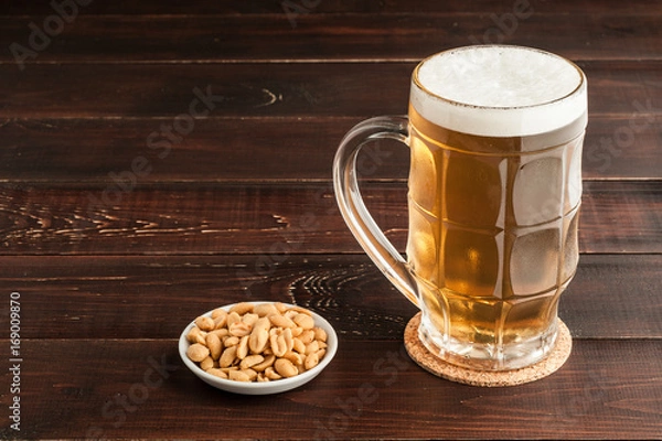 Fototapeta glass of cold frothy lager beer and peanuts plate on wooden table