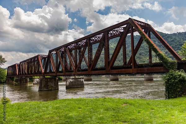 Fototapeta Old rusted railroad bridge crossing a river with green grass, mountains in the distance and a cloudy blue sky overhead.
