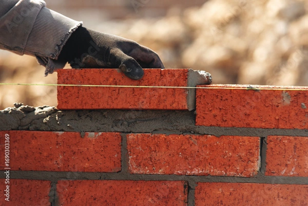 Fototapeta Skilled worker laying red bricks in a construction site during daylight with focused attention on building a wall