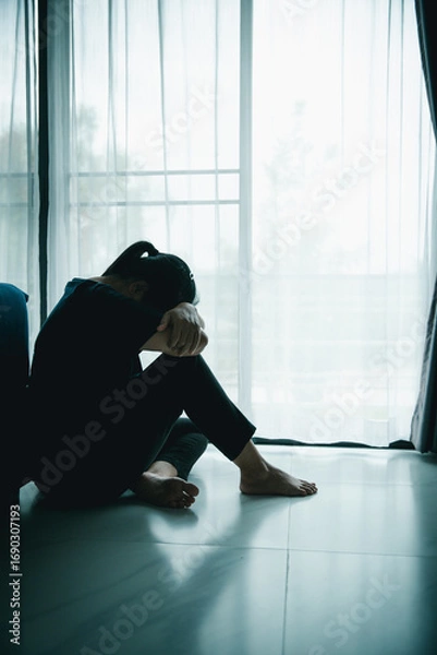 Fototapeta A lonely woman sits on the floor by window with hand on head in dark room. Concept of depression, sadness, anxiety, stress, mental health problem, loneliness and emotional struggle in life.