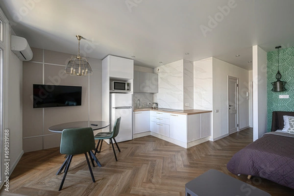 Fototapeta modern studio apartment featuring a small kitchen, dining area with green chairs, and a bed in a partitioned space. The interior design is clean and functional