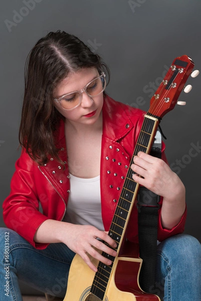Fototapeta Female guitarist and musician in process of tuning her acoustic guitar, holding it upright and plucking strings in order to ascertain sound of her preferred musical instrument