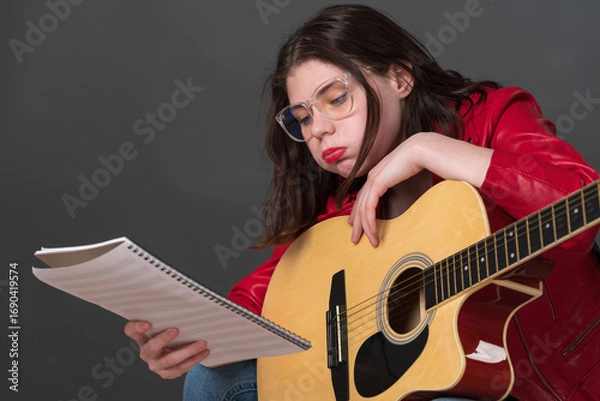 Fototapeta Female musician, equipped with guitar, meticulously examines sheet music while exhibiting countenance indicative of exhaustion during protracted and arduous rehearsal
