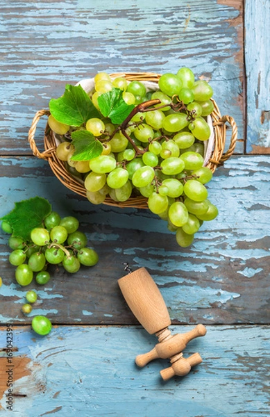 Fototapeta Overhead green grapes in knitted basket on old blue rustic wooden table and cork opener