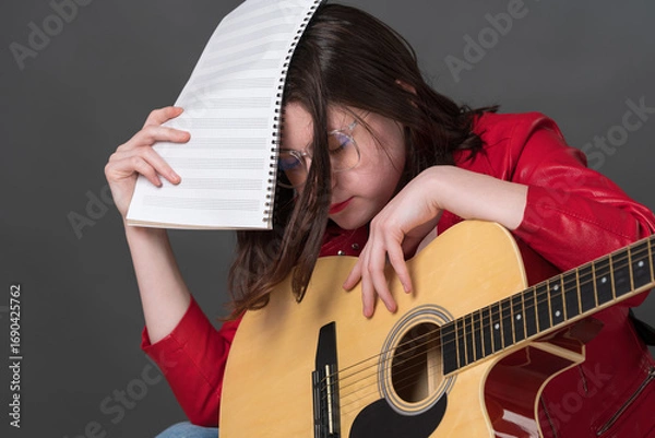 Fototapeta Tired and exhausted female musician with guitar and notes in hand covers face with music sheets due to fatigue from long and tiring rehearsal and learning to play classical six string acoustic guitar