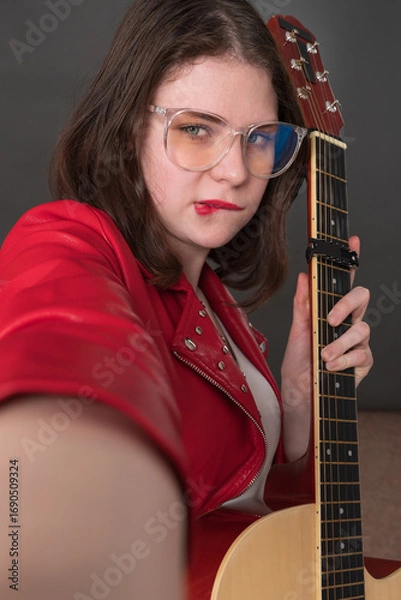 Fototapeta Female influencer guitarist biting her lip in playful expression takes selfie of herself with guitar in her hand, creating user-generated content