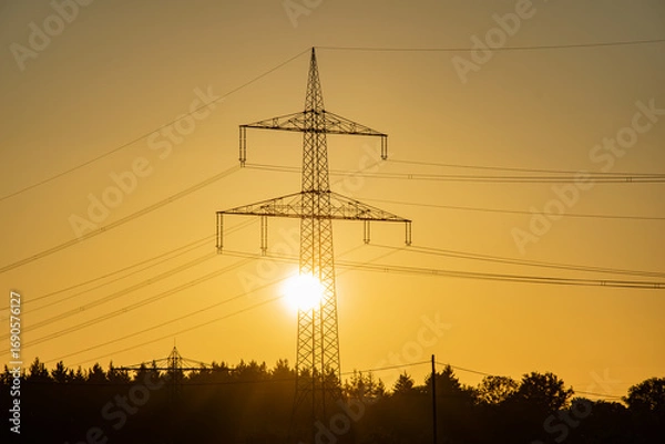 Fototapeta Transmission tower or high-voltage electricity pylon with sun shining through steel frame at sunset. Symbol of energy and power generation.