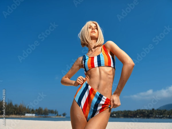 Fototapeta confident young woman wearing colorful striped bikini standing on sunny beach, posing with hands on hips, clear blue sky and distant trees in background