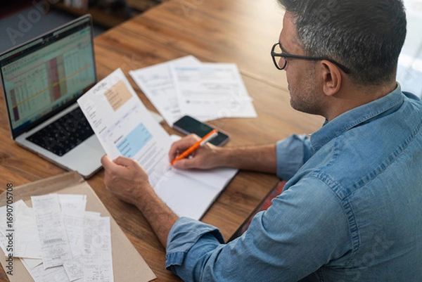 Fototapeta Mature man with glasses holds utility bill writing in paper notebook at laptop, male accountant calculates expenditures for company office service works, financial reports