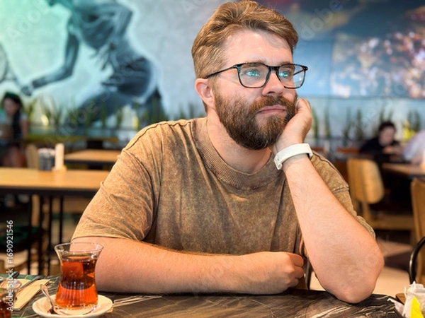 Fototapeta Bearded man with glasses sitting at cafe table with tea glass and thoughtful expression. Lifestyle, leisure, and indoor portrait representing social moment and casual dining.