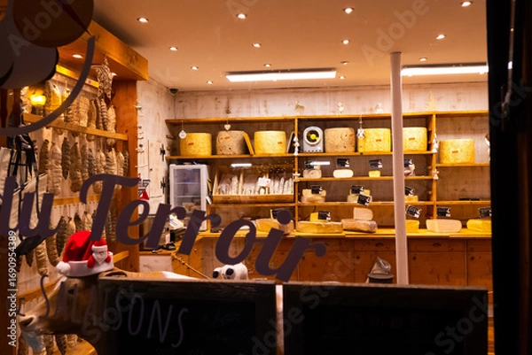 Fototapeta A cozy cheese shop showcasing an array of cheeses, wooden shelves filled with products, and festive holiday decorations in the evening ambiance.