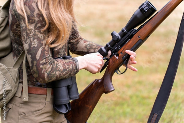 Fototapeta Female hunter checking her firearm with focus, dressed in full camouflage in natural landscape. Women in hunting with gun. Pre-hunt routine. Wilderness survival skills. Hunting culture and traditions