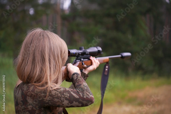 Fototapeta Woman in camouflage aims scoped rifle with concentration, standing in natural surroundings. Hunting in action. Female strength and wilderness survival skills
