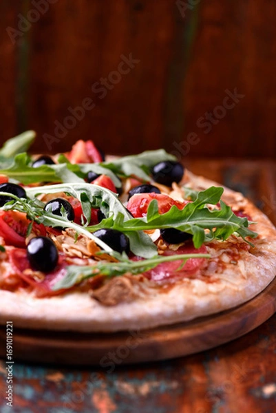 Fototapeta Pizza with salami, tomatoes and mozzarella cheese, black olives and arugula. Rustic wooden background. Soft focus. Copy space.	