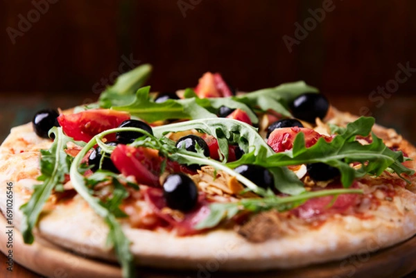 Fototapeta Pizza with salami, tomatoes and mozzarella cheese, black olives and arugula. Rustic wooden background. Soft focus. Copy space.	