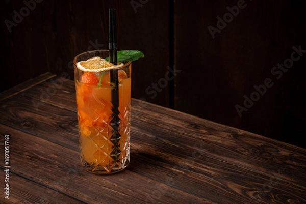 Fototapeta Refreshing highball cocktail with orange juice, basil, ice cubes and dried orange slice in tall crystal glass on wooden table