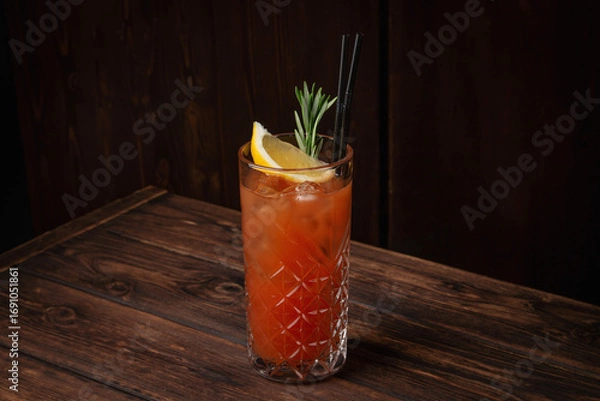 Fototapeta Classic tequila sunrise cocktail with lemon wedge, rosemary sprig and ice cubes in tall crystal glass on rustic wooden table