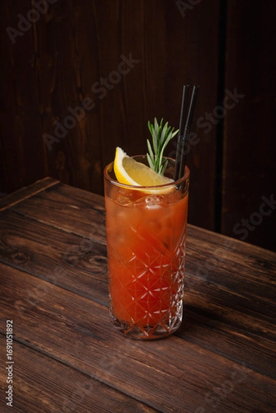 Fototapeta Classic tequila sunrise cocktail with lemon wedge, rosemary sprig and ice cubes in tall crystal glass on rustic wooden table