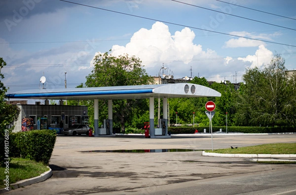 Fototapeta An everyday scene of a gas station with a "no entry" sign and a parked car under a dramatic cloudy sky. The puddles on the pavement suggest recent rain, creating a quiet, moody urban landscape.

