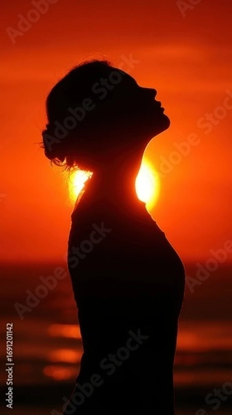 Fototapeta A woman is standing on the beach, looking up at the sun. The sun is setting, casting a warm glow over the scene. The woman is in a contemplative or meditative state