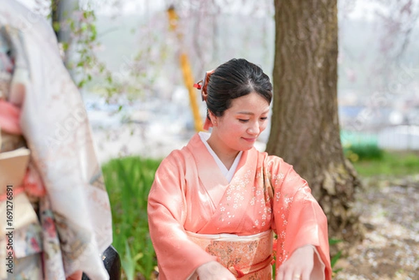 Fototapeta A woman in a traditional Japanese kimono sits under a tree. She is smiling and she is enjoying the peaceful surroundings