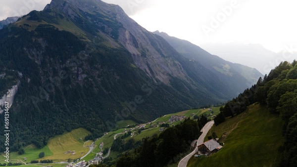 Fototapeta Swiss Alps with houses, winding road, clouds, and overcast sky, captured by drone. Schweizer Alpen mit Häusern, kurviger Straße, Wolken und bedecktem Himmel, aufgenommen mit Drohne.