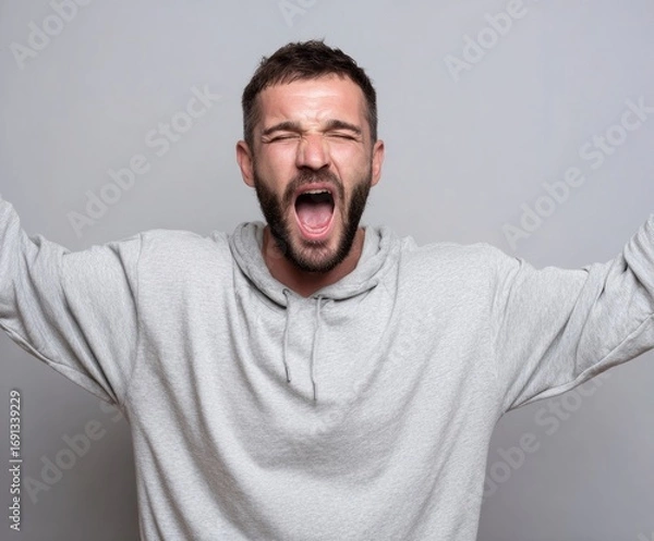 Fototapeta A man in a light gray hoodie expresses intense emotion with his mouth wide open in a dramatic, expressive portrait.