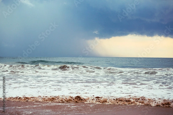Fototapeta Ocean waves rolling onto sandy beach with storm clouds on horizon