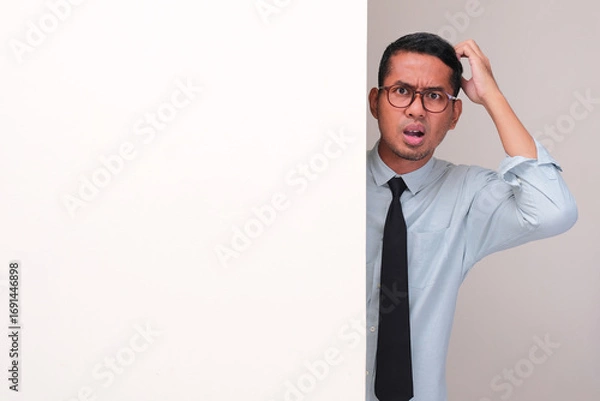 Fototapeta A company employee peeking behind wall scratching his head showing confused face expression