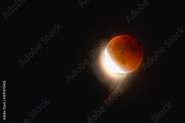 Fototapeta Lunar Eclipse on Sunday 7 September, 2025. The Moon emerges from the Earth's shadow, and causes an ethereak effect on high altitude cirrus clouds.