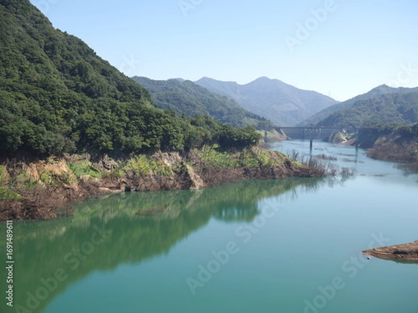 Fototapeta Gunma, Japan - September 7, 2025: Appeared due to drought, submerged disused railroad tracks, roads, basements and trees under the water of Yamba Dam in Gunma, Japan