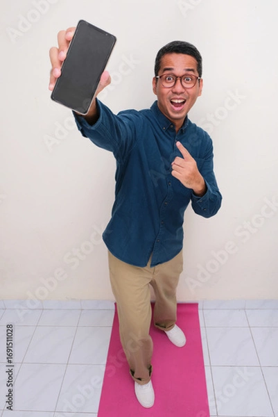 Fototapeta High angle view of adult asian man standing while showing blank phone display screen with amazed face