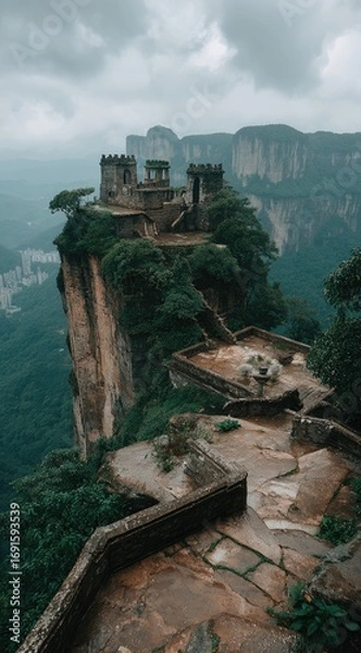Fototapeta An ancient stone fortress perched atop a misty cliff overlooking lush valleys and distant mountains on a cloudy day