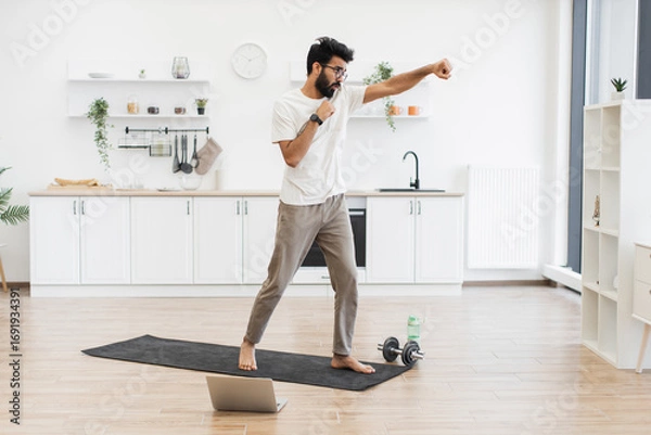 Fototapeta Man in 30s practicing boxing exercises in spacious kitchen on yoga mat, showing fitness.